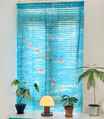 Decorative window blind with fish design on a white wall with plants and a lamp below.