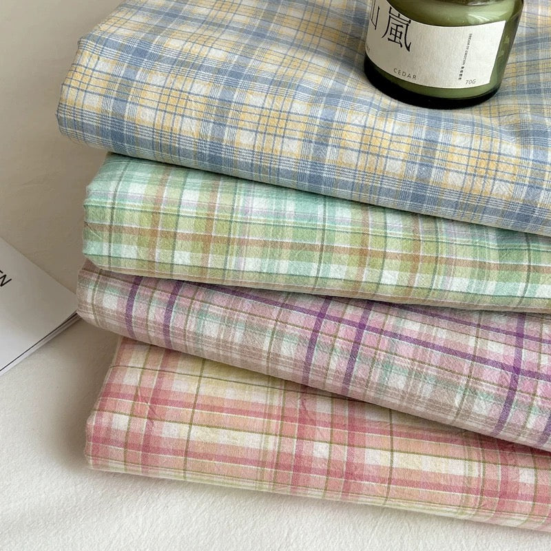 Four folded plaid fabric squares stacked on a light surface, each showing a different pastel plaid pattern: blue and yellow, mint and peach, pink and purple, and coral and cream. A dark green candle jar sits on the top folded fabric.