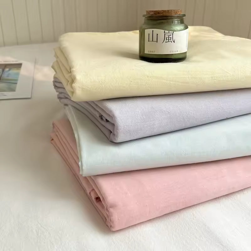 Four folded fabric sheets or linens in pale cream, lavender, mint, and blush pink are stacked on a white surface, with a small glass jar candle on top. No person or real environment is meaningfully present beyond a simple styled tabletop setup.