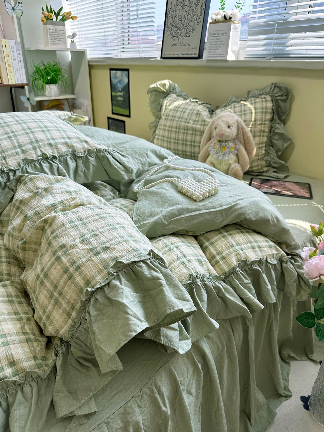 Bed with green plaid bedding, a teddy bear, and flowers in a room with a window and plants.