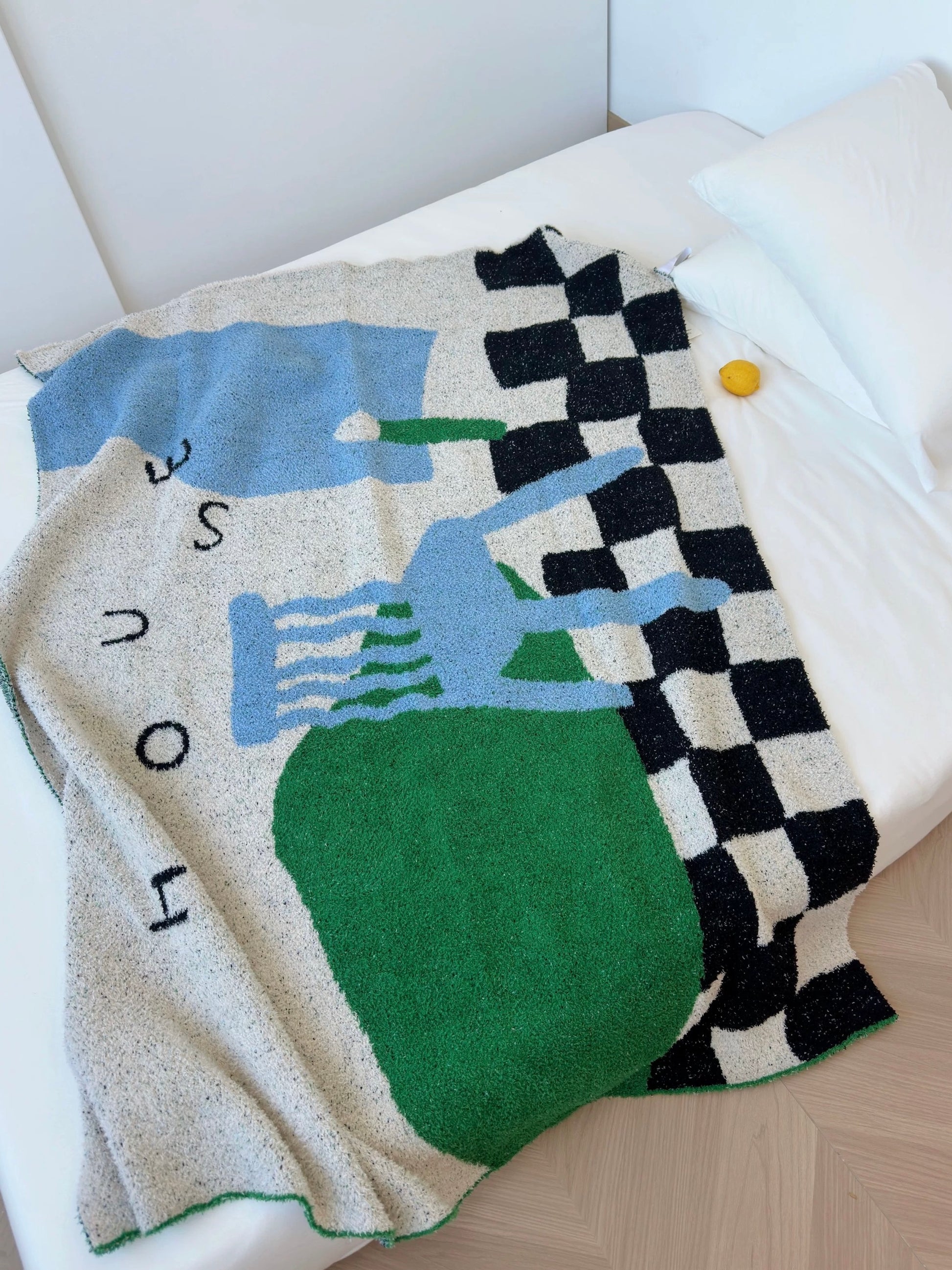 A textured knit throw blanket draped over a white bed. The blanket features a modern abstract design with a large green shape, light blue shapes, a black and white checkerboard pattern, and the word 'HOUSE' in black letters on a speckled cream background.