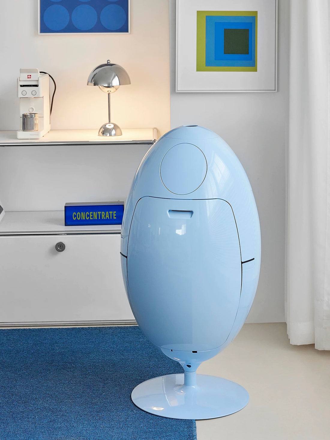 Blue egg-shaped chair in a room with a window and artwork on the wall.