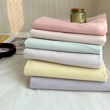 A stack of six folded cotton fabrics in pastel shades of pink, cream, mint, light purple, pale yellow, and lavender, arranged on a white surface with a small glass jar on top.