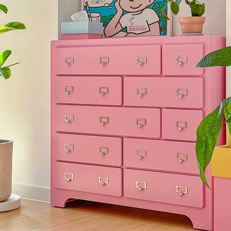 Pink dresser with multiple drawers in a room setting with plants and a poster.