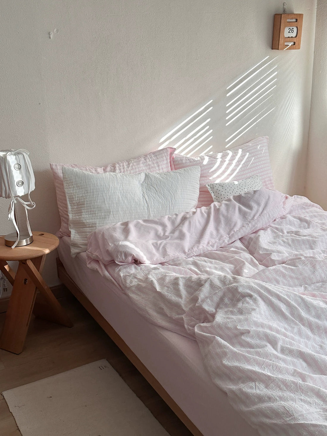 A bed dressed in light pink and white striped bedding, including a duvet cover, matching pillowcases, and several pillows, set in a bedroom with a wooden side table and a small wall clock.