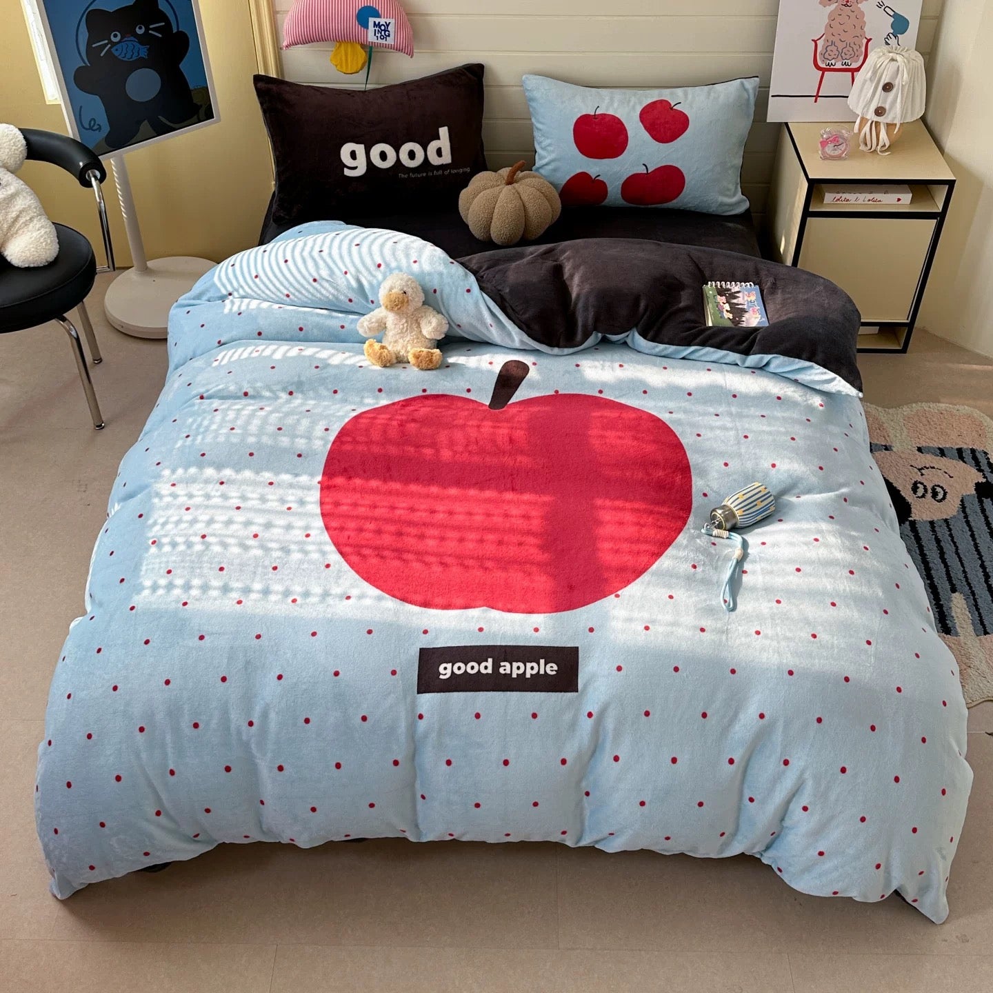 Bed with blue comforter featuring a red apple design and 'good apple' text, surrounded by toys and pillows.