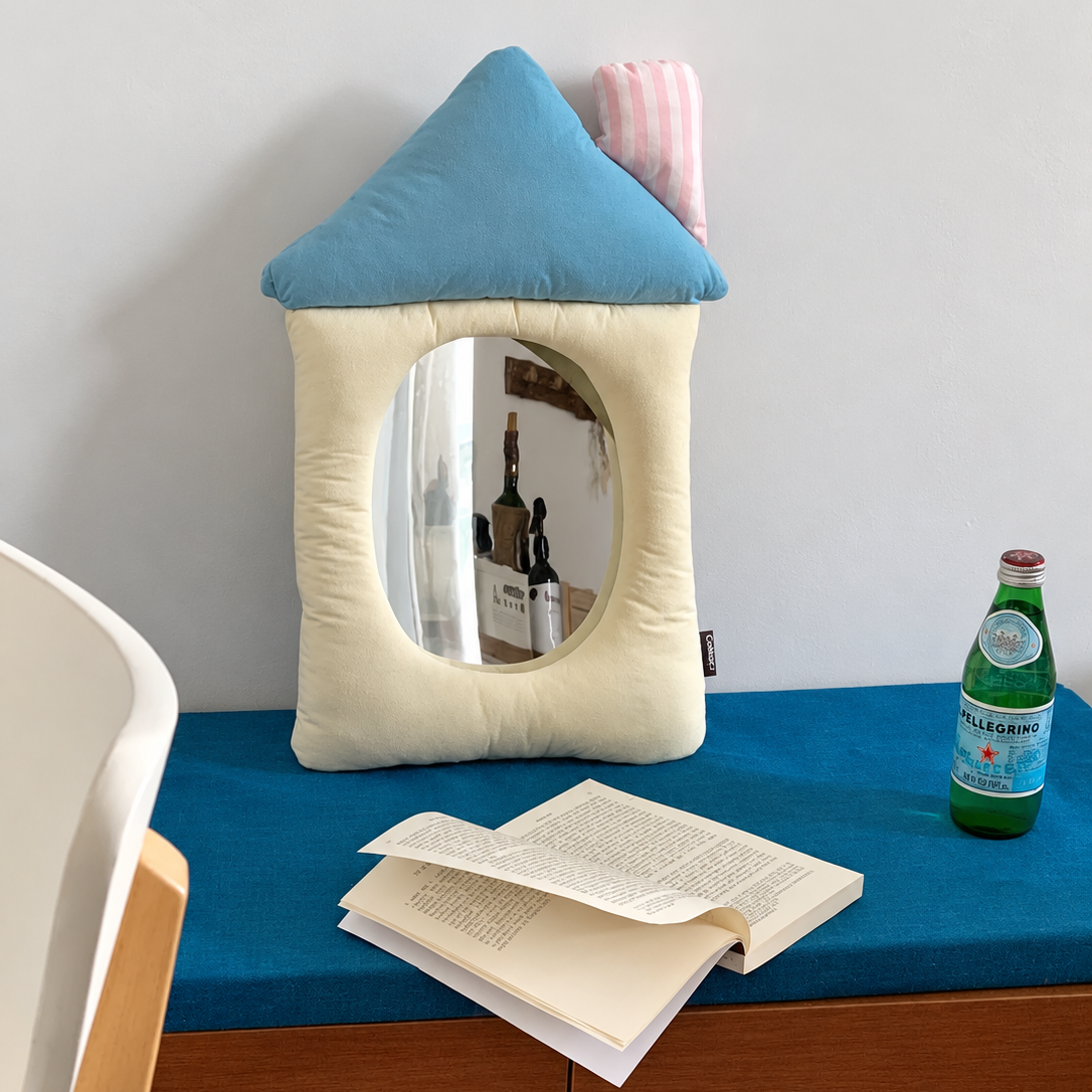 A house-shaped plush cushion mirror with a blue roof and cream body, featuring an oval mirror. It is styled on a blue-clothed wooden cabinet next to an open book, a green San Pellegrino bottle, and a pink striped pillow against a plain white wall.