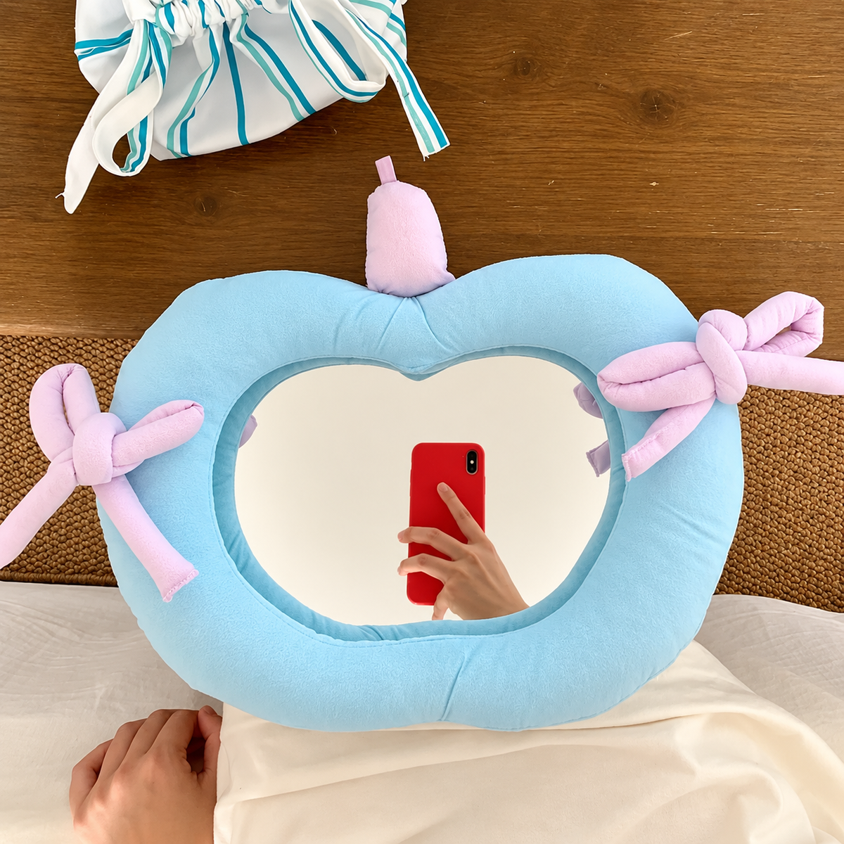 A light blue heart-shaped plush pillow with a mirror inside and pastel pink knotted bow details, shown on a bed with a person taking a mirror selfie. A matching blue and white striped drawstring bag is visible in the background on a wooden surface.