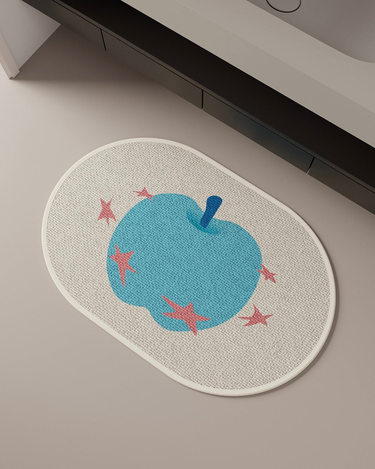 An oval bath mat with a textured beige woven background, featuring a large blue apple design with pink starfish accents and a blue handle, shown on a neutral bathroom floor near a bathtub.