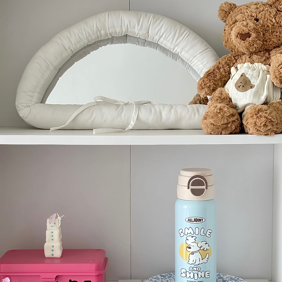 A light beige padded baby crescent-shaped mirror with a white ribbon bow, displayed on a white shelf next to a brown teddy bear. Below the shelf, a light blue Alljoni water bottle is visible.