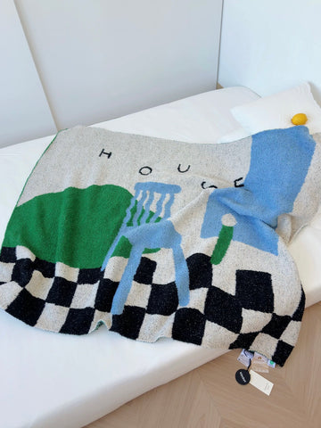 A textured knit throw blanket with a colorful abstract design and a black-and-white checkered border, draped over a white bed. The design features green, light blue, and gray blocks with the word 'HOUSE' printed above.