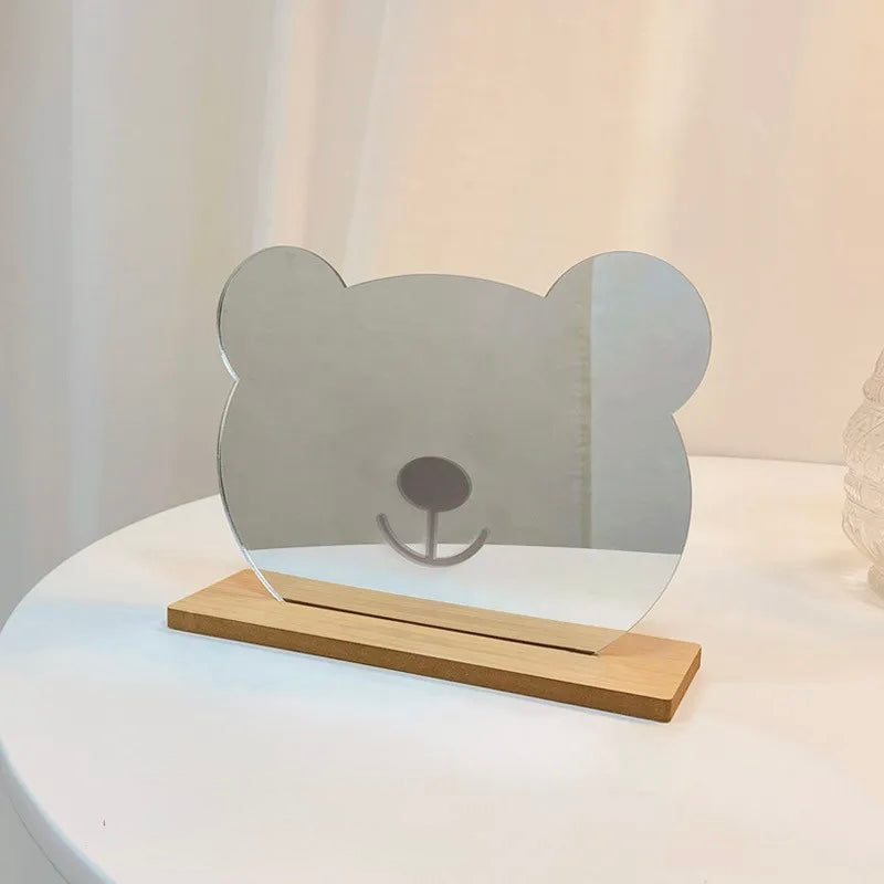 A small tabletop mirror shaped like a bear head, mounted on a light wood base, displayed on a white round table against a soft cream background.