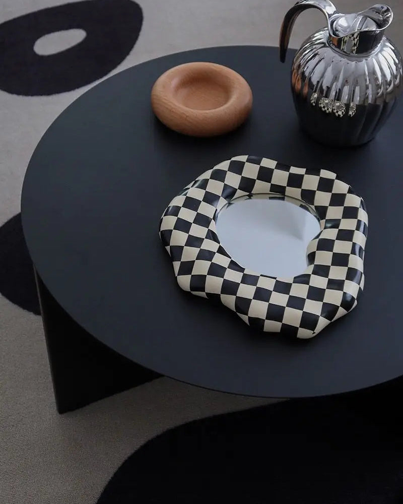 A black and white checkered cushioned mirror with a scalloped edge, styled on a round black coffee table next to a wooden bowl and a silver pitcher.