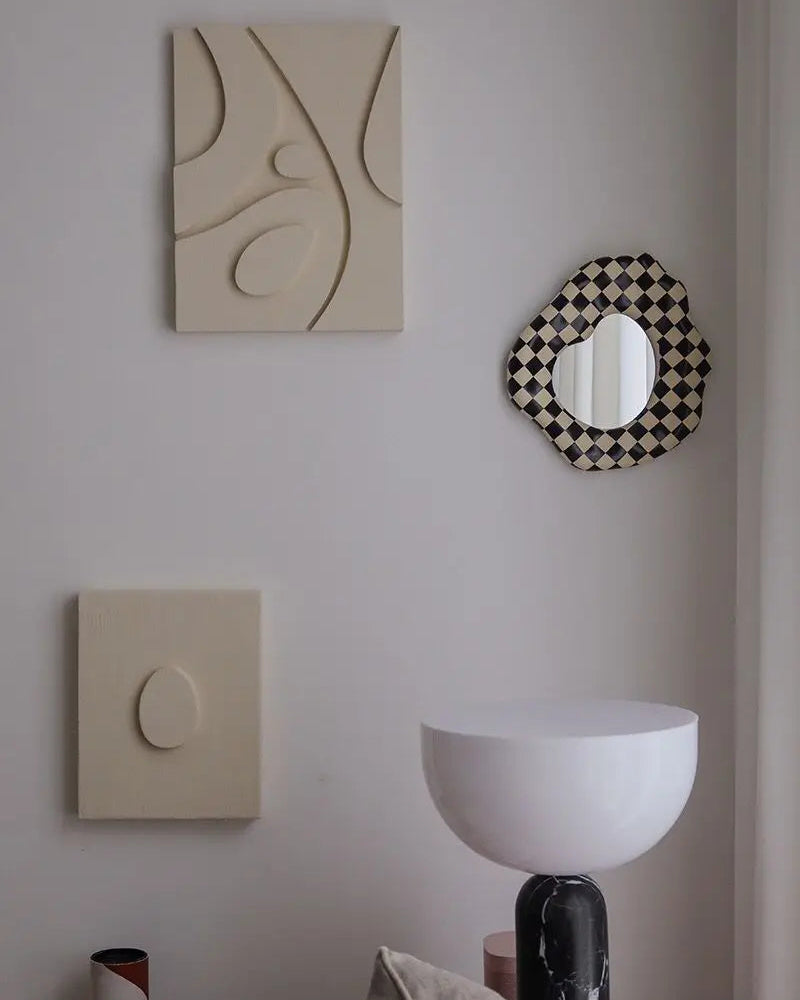 A styled interior wall with abstract wall art in warm cream tones, a black-and-white checkered scalloped mirror, and a white bowl-shaped table above.