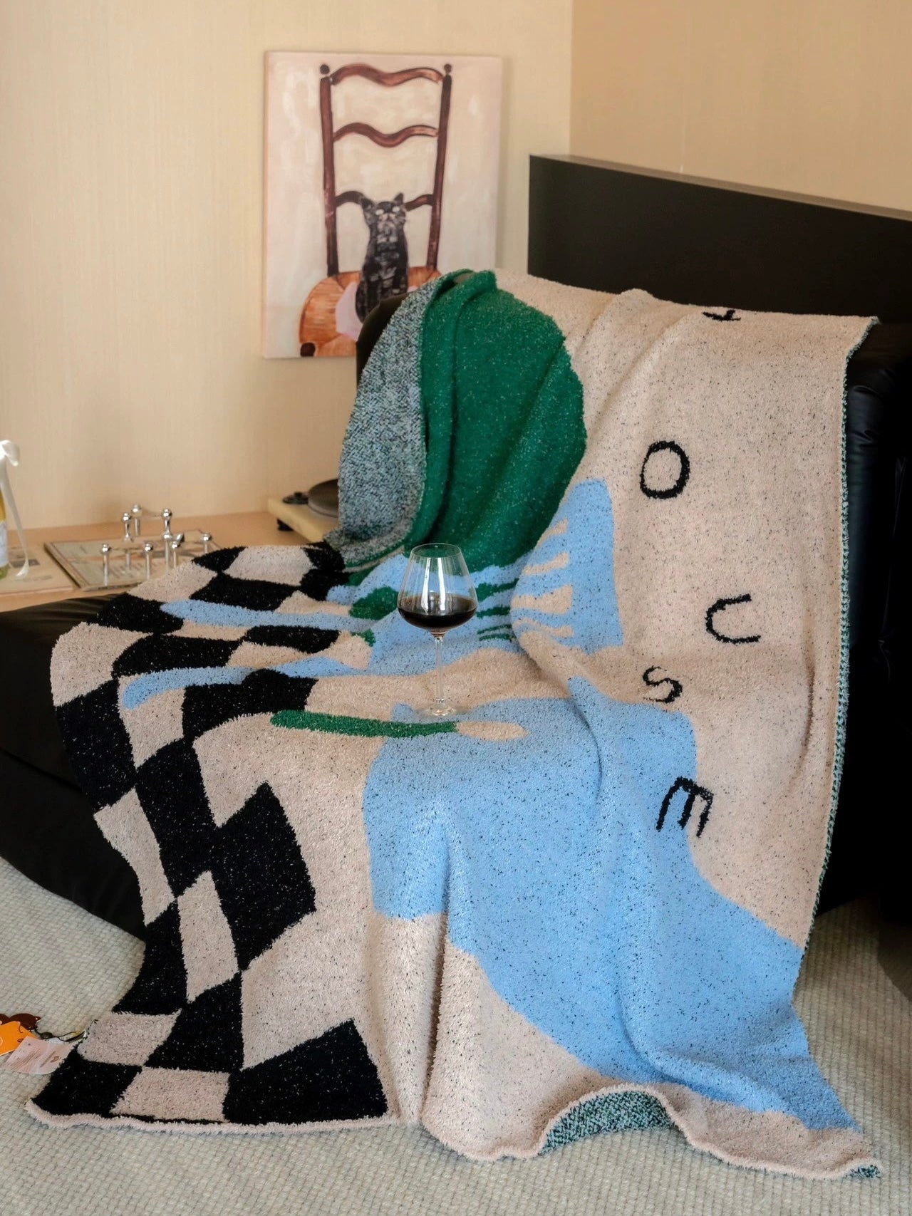 A chunky knit throw blanket draped over a dark armchair in a bedroom setting. The blanket features a modern abstract design with black, light blue, forest green, and beige sections, including a black-and-white checkerboard pattern on one side and stylized lettering on the light blue section.