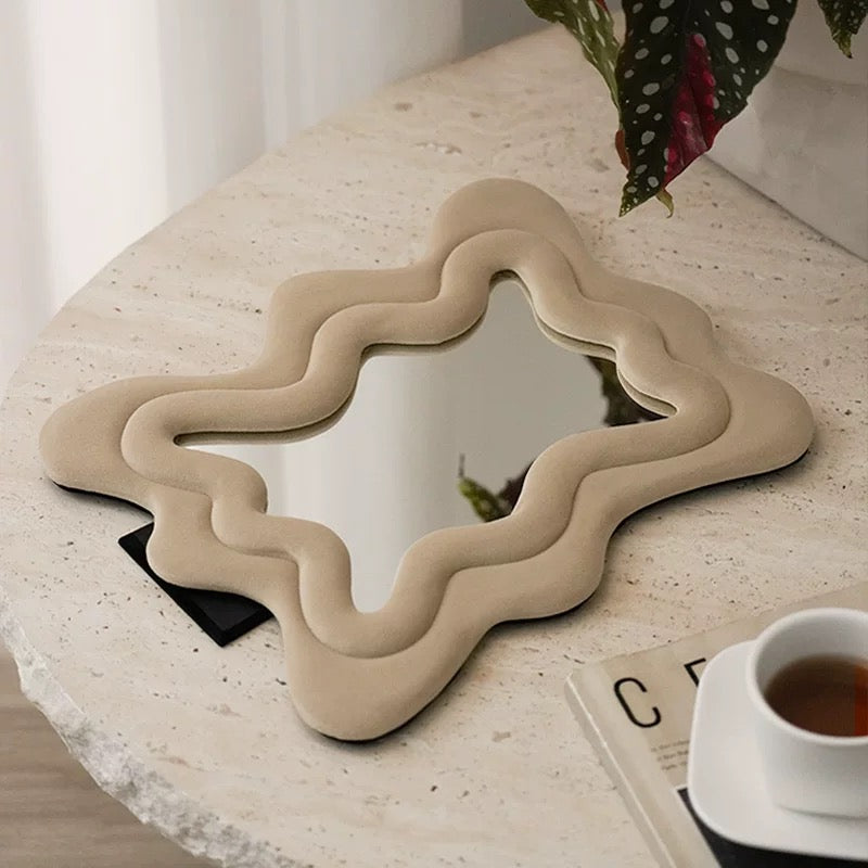 A wavy, multi-tiered beige mirror with a thick sculptural frame, styled on a textured stone tabletop next to a cup of tea and a plant.