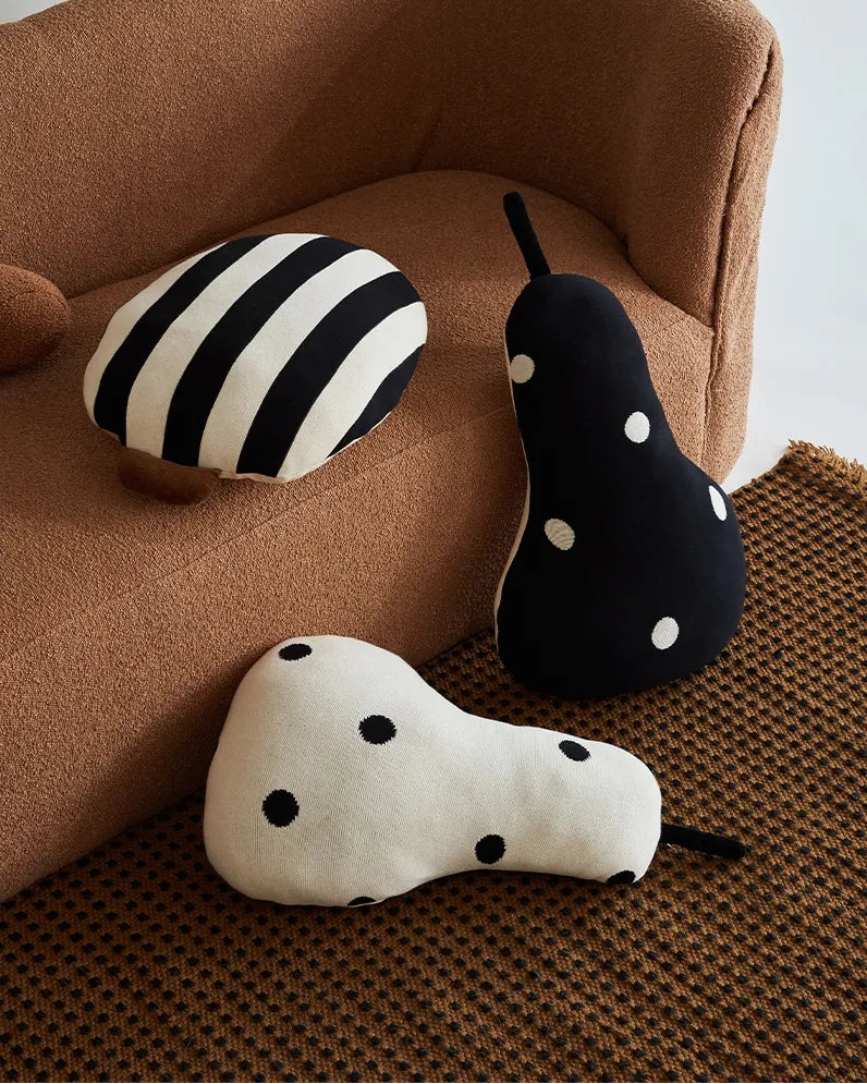 Three decorative throw pillows styled in a home setting: a pear-shaped pillow with black polka dots on an off-white background, a black and white striped oval pillow, and a dark navy pear-shaped pillow with white polka dots. They are arranged on a textured brown rug next to a brown upholstered armchair.
