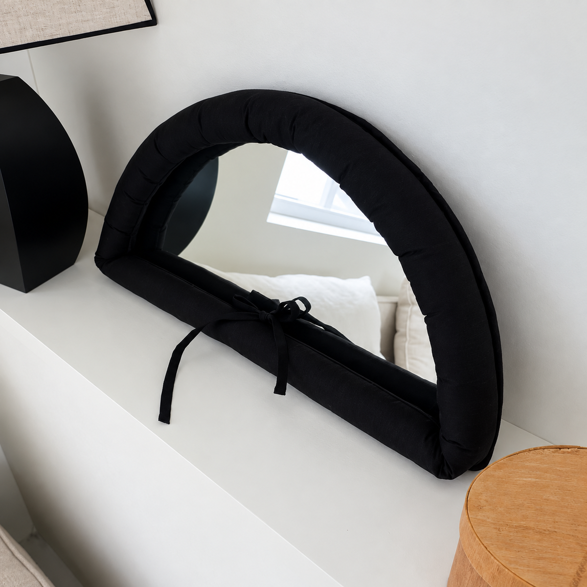 Black half moon mirror with a gathered fabric frame and matching ribbon bow, propped against a pale wall.