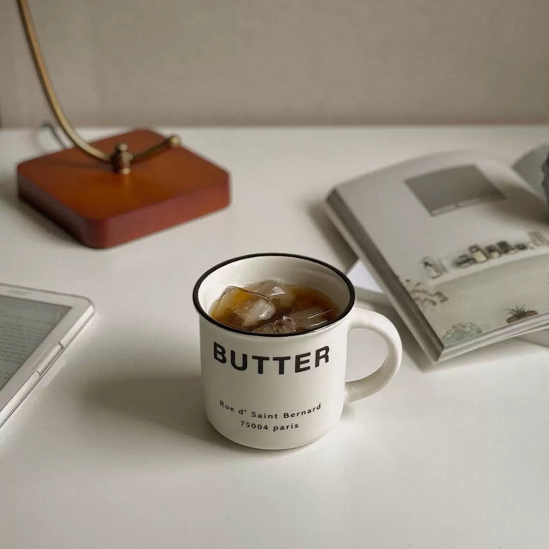 A white ceramic mug with a black rim and the word 'BUTTER' printed on it, filled with iced coffee, sitting on a white desk next to an open book and a lamp.