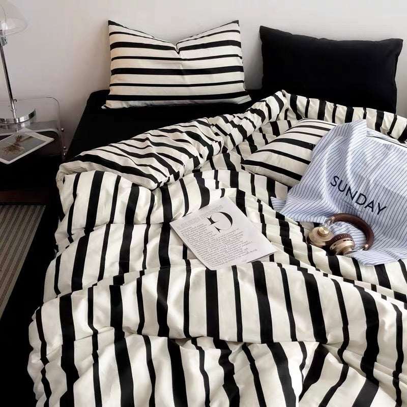 A bed styled with a black and white striped bedding set, including a duvet cover and matching pillowcases, alongside a solid black pillow. A blue-and-white striped pillowcase with the word 'SUNDAY' is also visible, along with headphones, a book, and a bedside lamp in a bedroom setting.