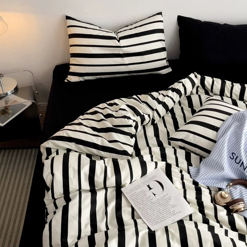 A cozy bed setup featuring a black and white striped bedding set, including a duvet cover and matching pillowcases, styled with a black pillow, a bedside table, and a magazine.