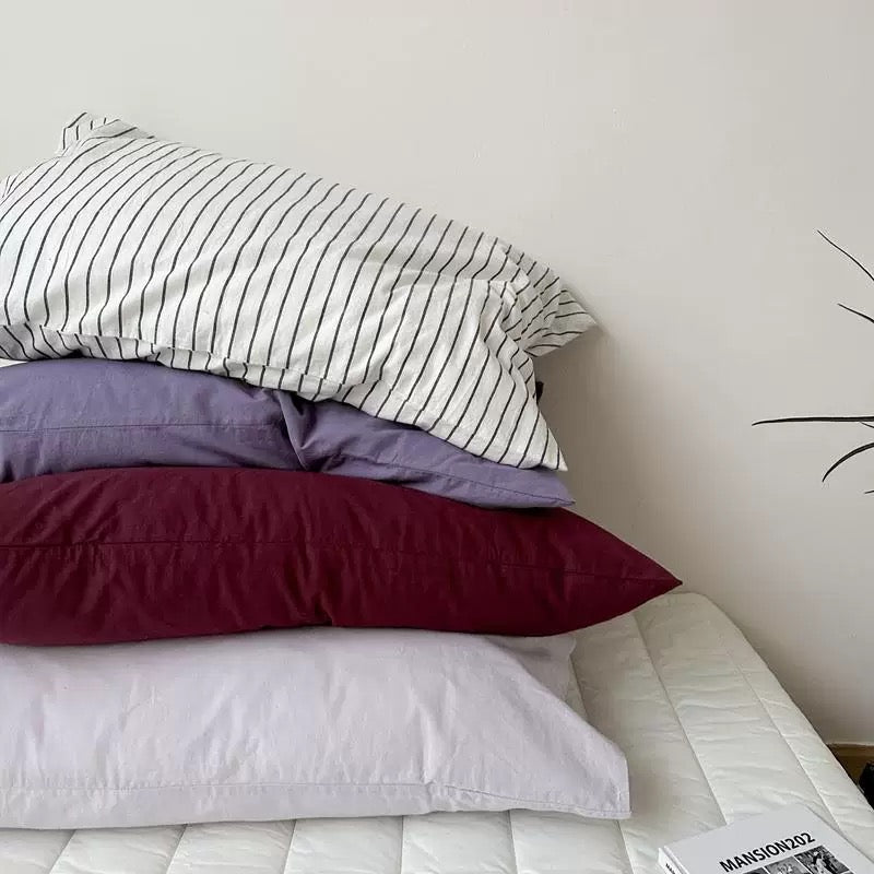 A lavender pillowcase is stacked with plum, pale gray, and white striped pillowcases on a white bed.