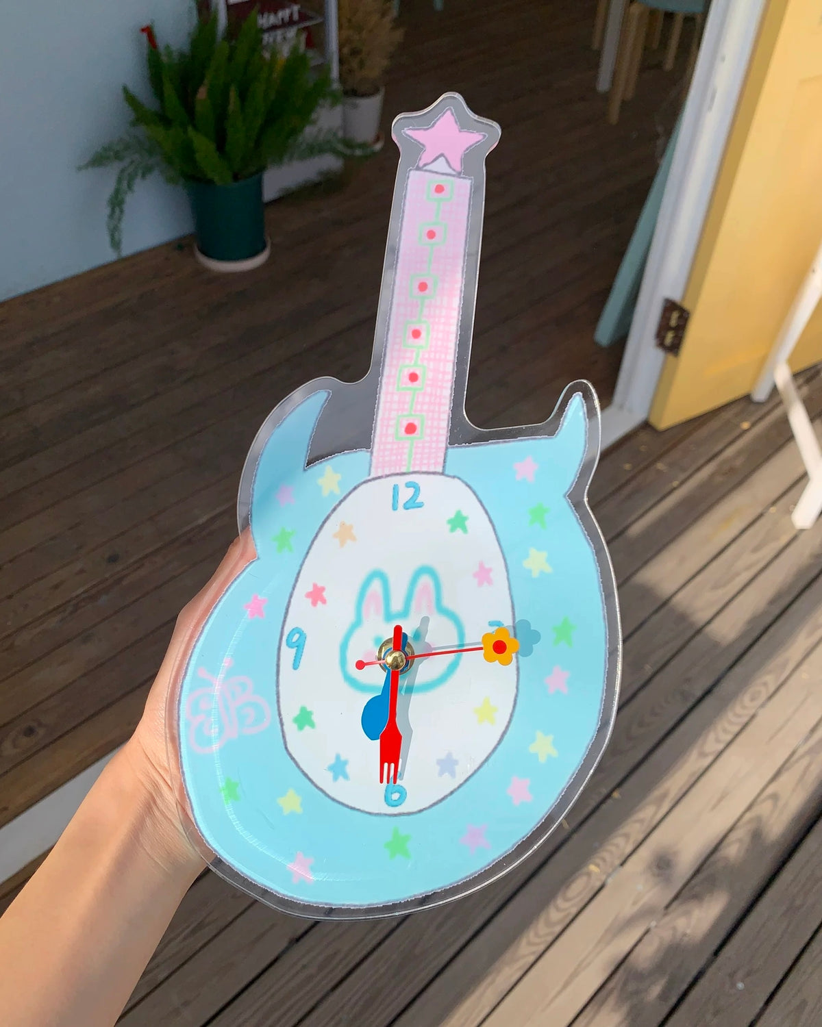 A hand holds a guitar-shaped clock outdoors on a wooden deck. The clock has a light blue body, a pink neck, a bunny face on the center, multicolored star accents, and red and blue hands.