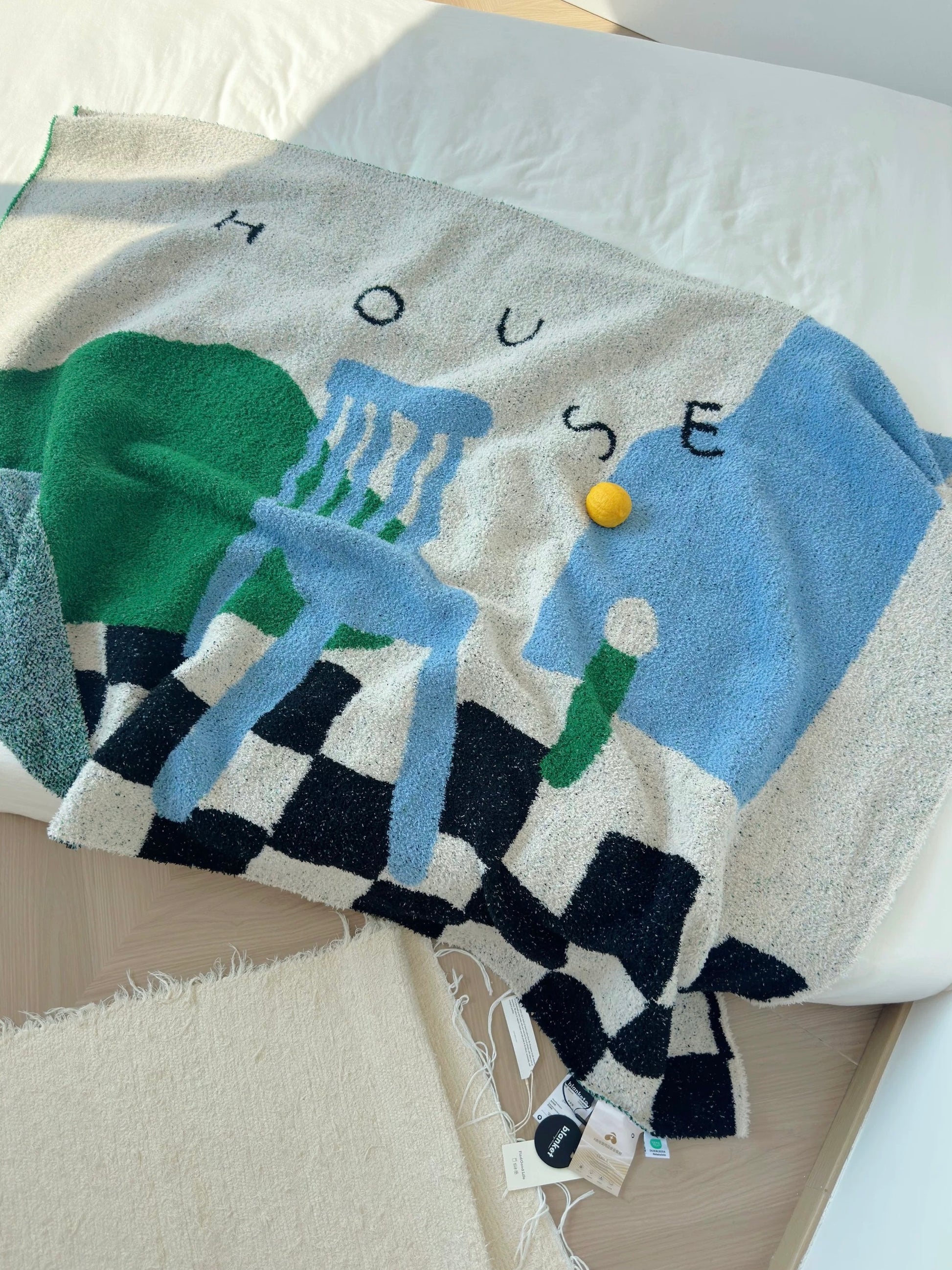 A chunky knit throw blanket draped over a bed, featuring a colorful abstract design with the word 'HOUSE' in black letters, a blue chair motif, green shapes, a black-and-white checkerboard pattern, and a small yellow accent.