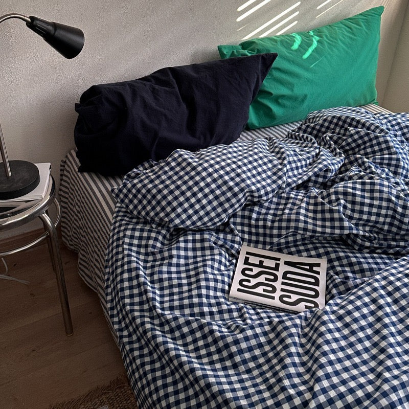 A bed in a real bedroom setting with a navy and white gingham duvet cover, a dark navy pillow, a green pillow, a black bedside lamp on a metal side table, and a magazine on the duvet.