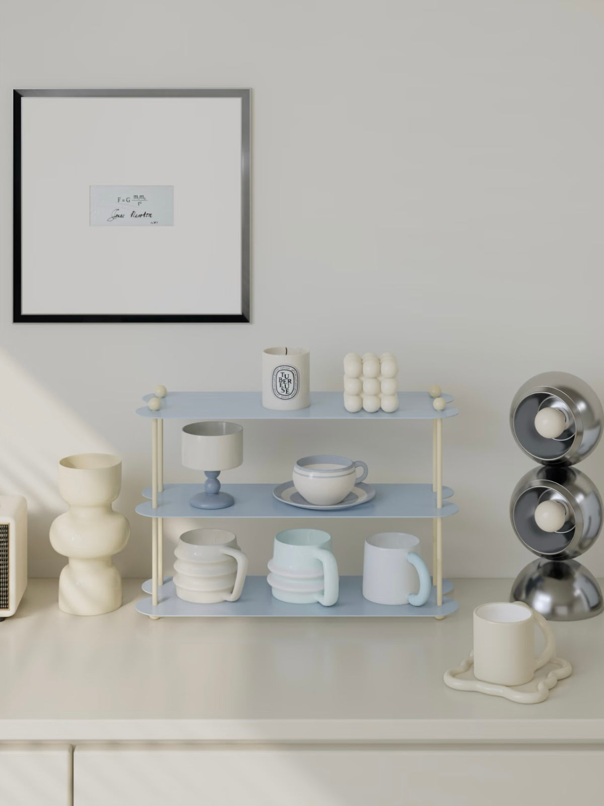 A blue display shelf sits below a framed print, with cups on its lower tiers and candles on the top tier.