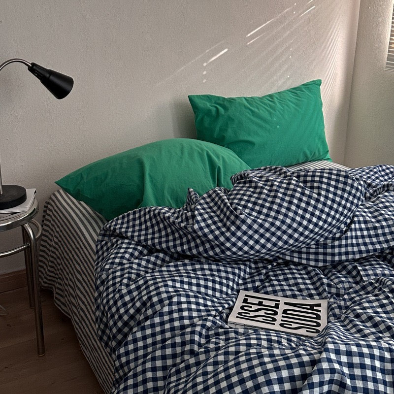 A bed styled in a bedroom setting with a navy and white gingham check duvet cover, matching pillowcases, and two solid green pillows. A black metal desk lamp sits on a side table next to the bed, and a book with the word 'STUDY' printed on it rests on the duvet.