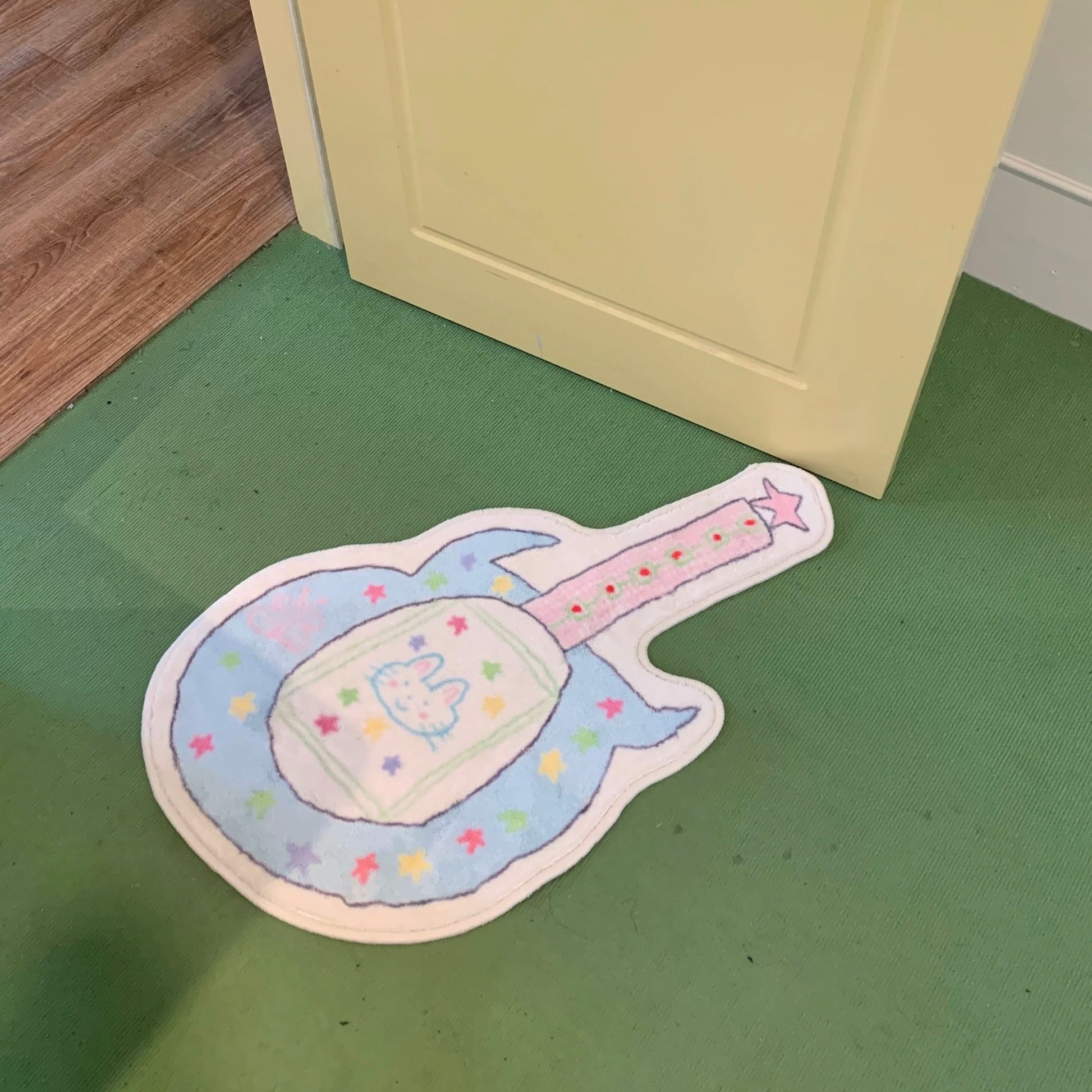 A guitar-shaped floor mat with a pastel blue body, pink neck, and colorful star details, placed on a green floor next to a cream-colored door.
