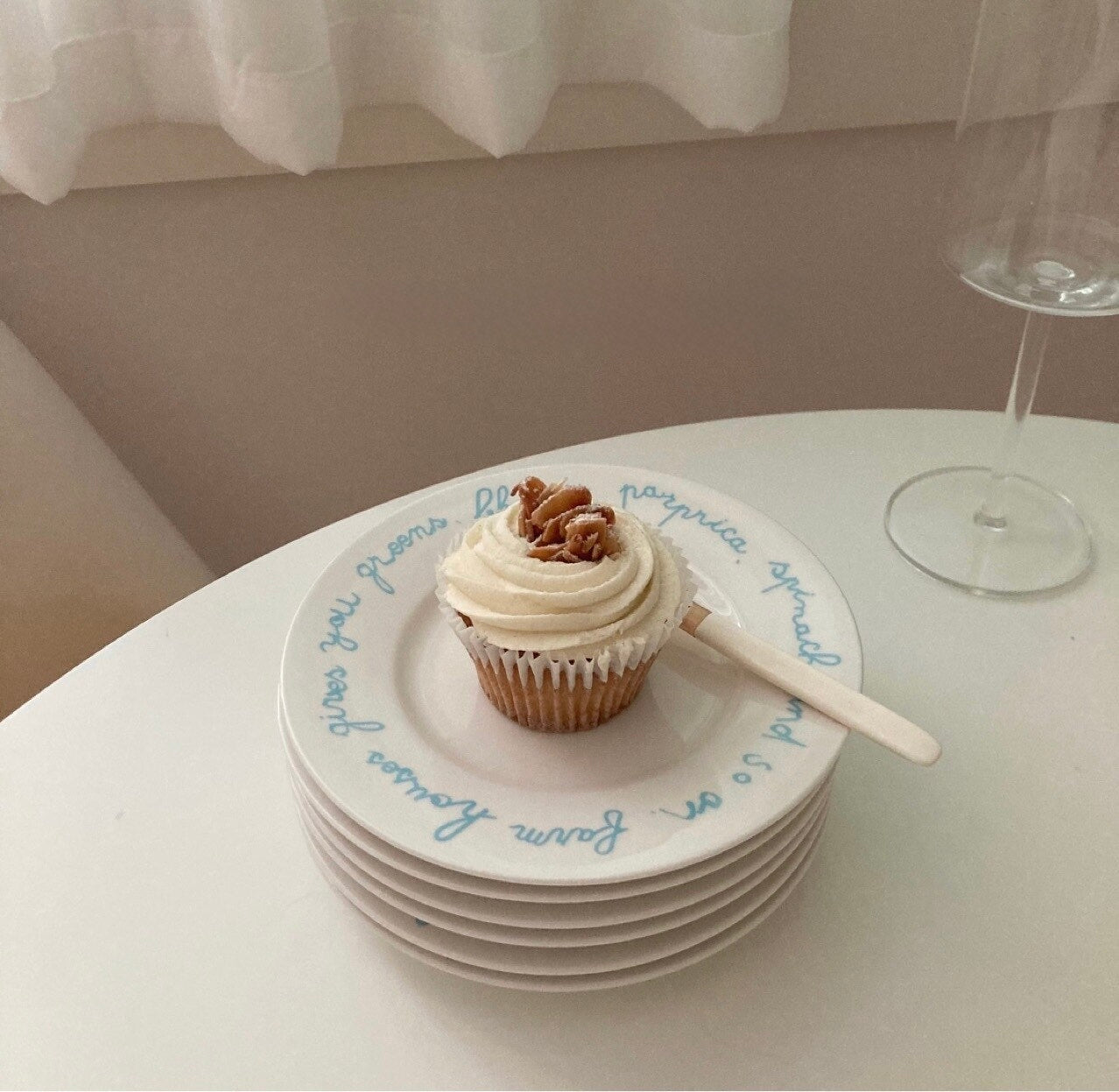 A stack of white ceramic plates with blue cursive writing, featuring a single frosted cupcake topped with walnuts and a wooden spoon, set on a white table next to a wine glass.