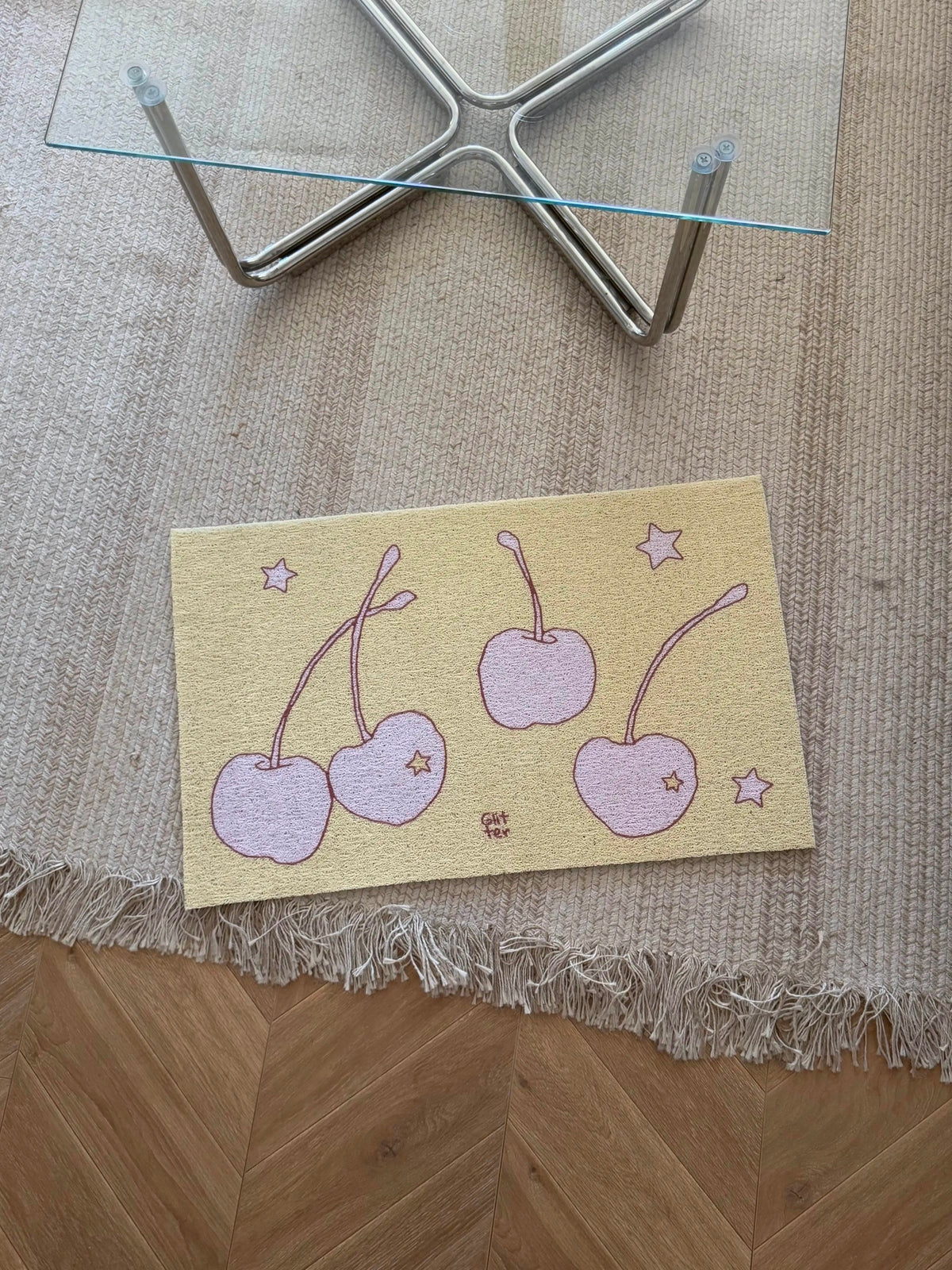 A rectangular decorative mat or tray with a pastel cherry illustration lies on a woven beige rug over a wood floor, with the lower legs of a glass-top table visible above.