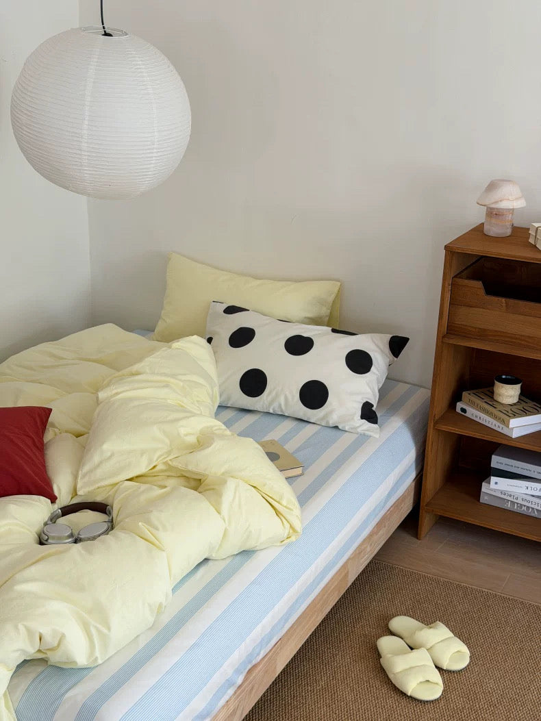 A cozy bedroom scene showing a bed with a pale yellow duvet, a light blue and white striped sheet, a black-and-white polka dot pillow, and a wooden nightstand beside it.