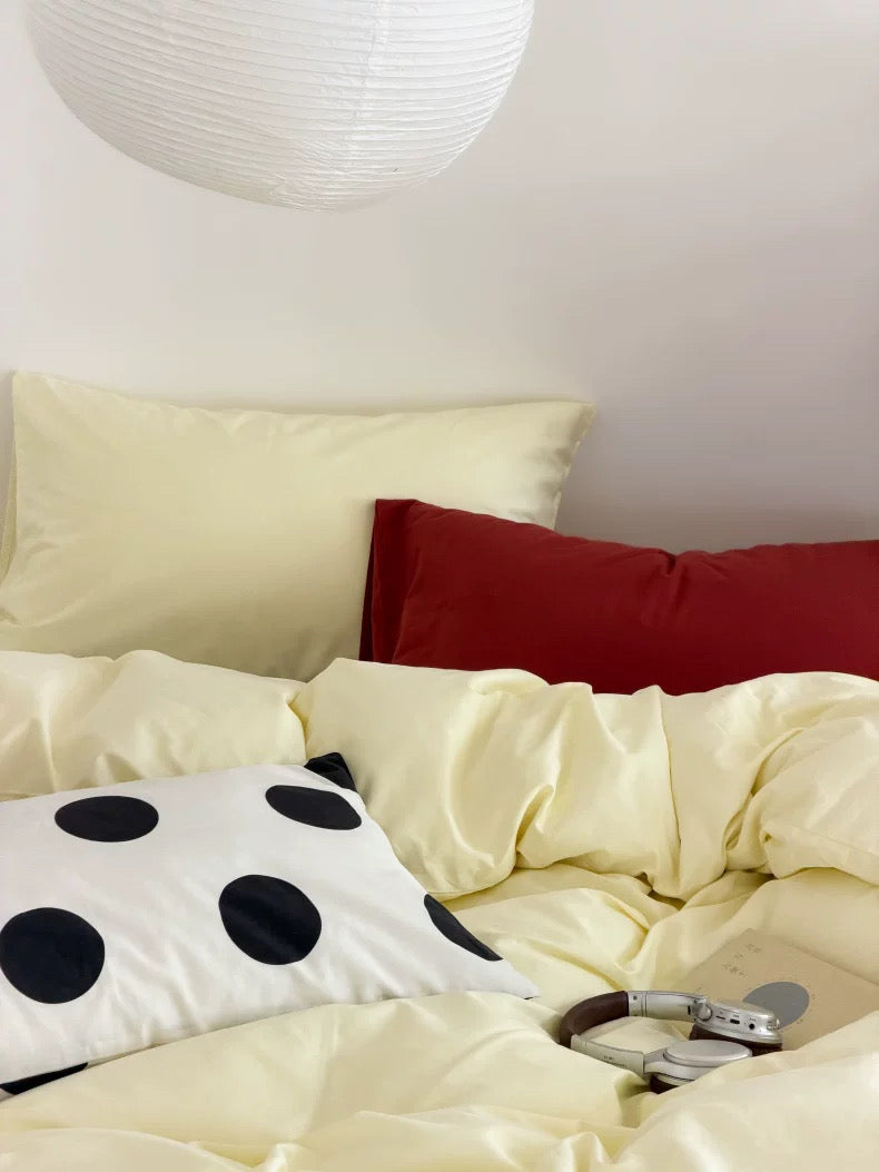 A cozy bed setup with cream-colored bedding, a red accent pillow, a white pillow with black polka dots, and a white hanging paper lantern above.