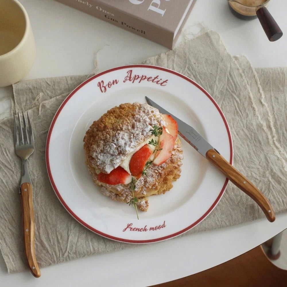 A white ceramic plate with red trim, printed with 'Bon Appetit' and 'French mood', holding a strawberry and cream cake. The plate is styled on a beige textured placemat with a wooden-handled knife, a fork, a cream mug, and a book in the background.