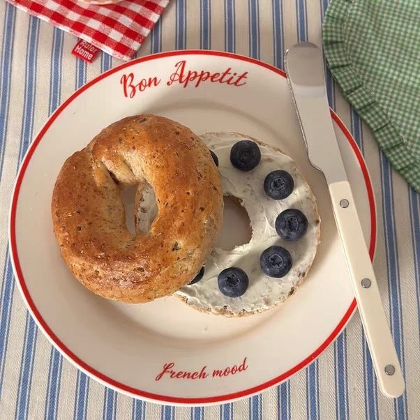 A white ceramic plate with red trim, printed with the words 'Bon Appetit' and 'French mood', holding a whole seeded bagel and a cut bagel spread with cream cheese and topped with blueberries. A metal spreader knife with a white handle rests on the right side of the plate, set on a blue and white striped tablecloth.