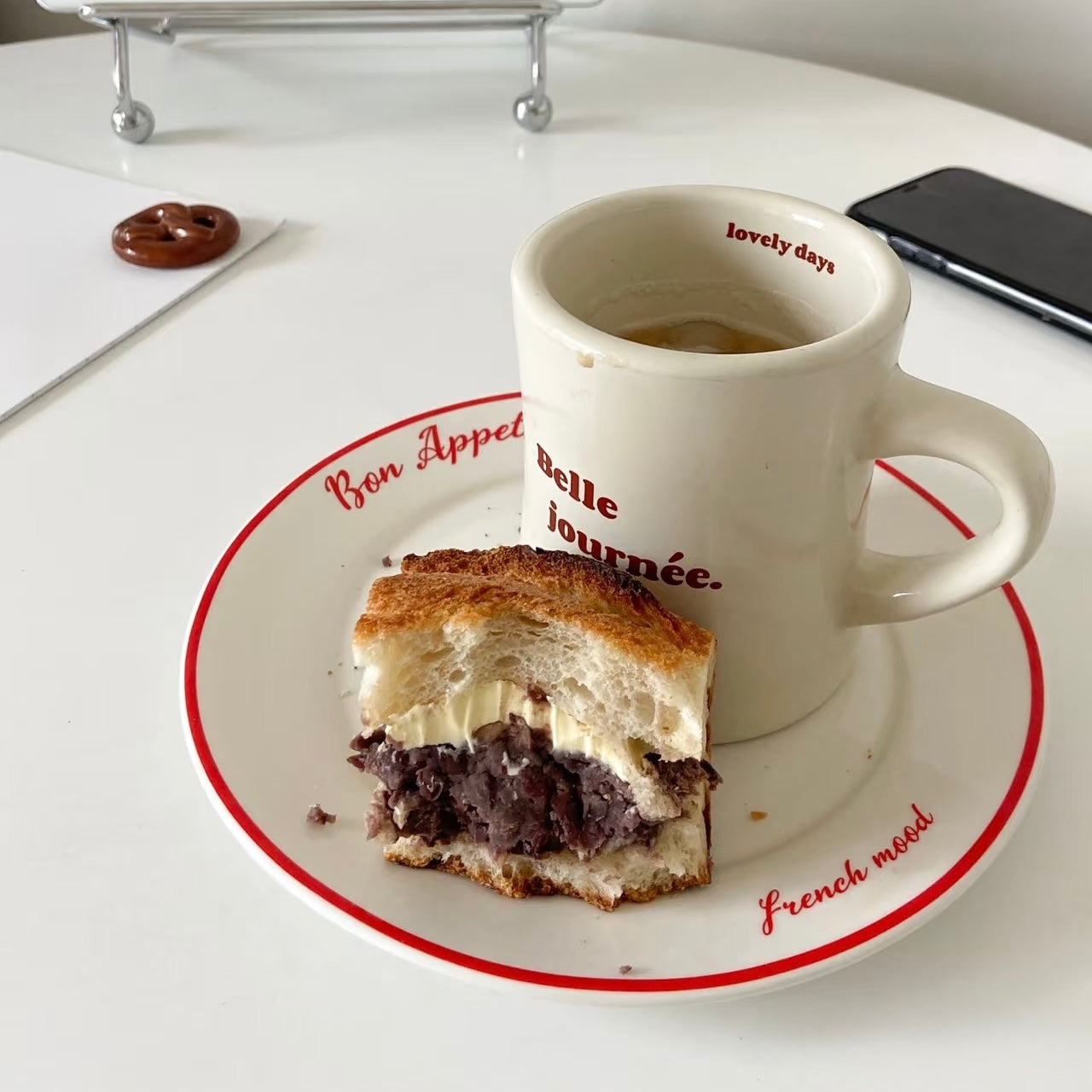 A white ceramic coffee mug with red text reading 'bon appét belle journée.' sits on a matching white plate with a red border, next to a partially eaten sandwich on a white tabletop.