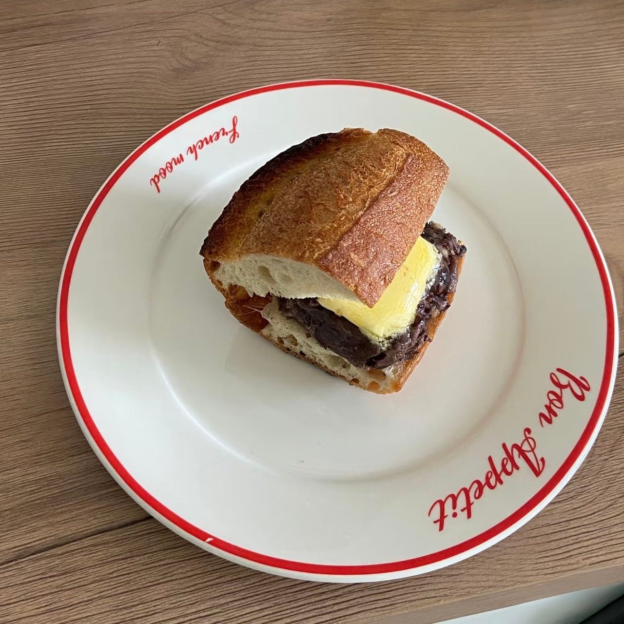 A close-up shot of a gourmet sandwich on a white ceramic plate with red trim, set on a wooden table. The sandwich features a rustic baguette, a thick beef patty, and melted yellow cheese.