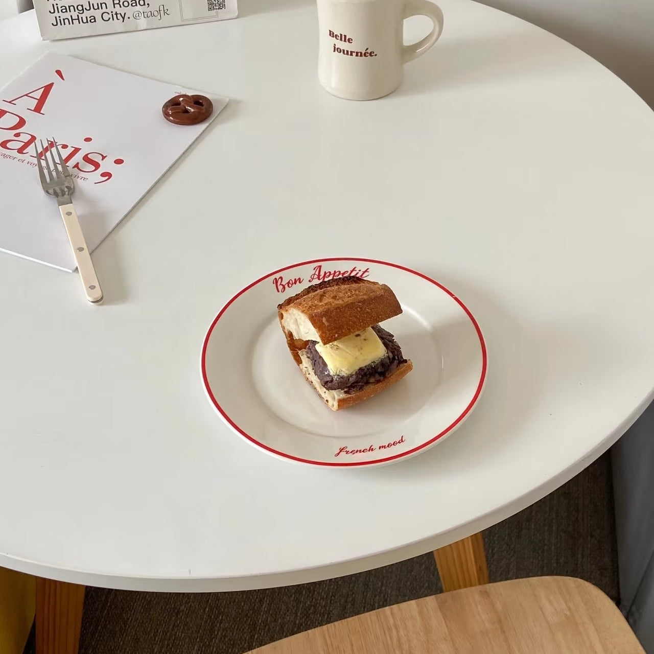 A small sandwich on a white plate with a red rim, set on a white round table with a mug, book, and fork nearby.