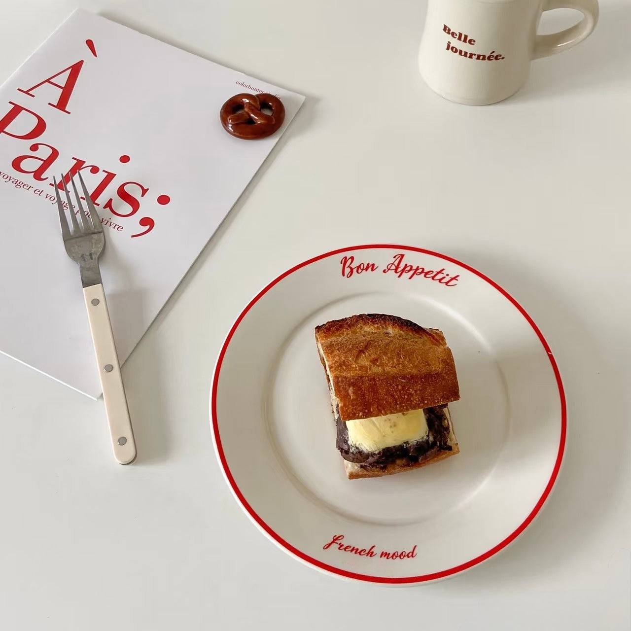 A small toasted sandwich on a white plate with a red rim, styled on a white tabletop with a fork, a magazine, and a mug nearby.