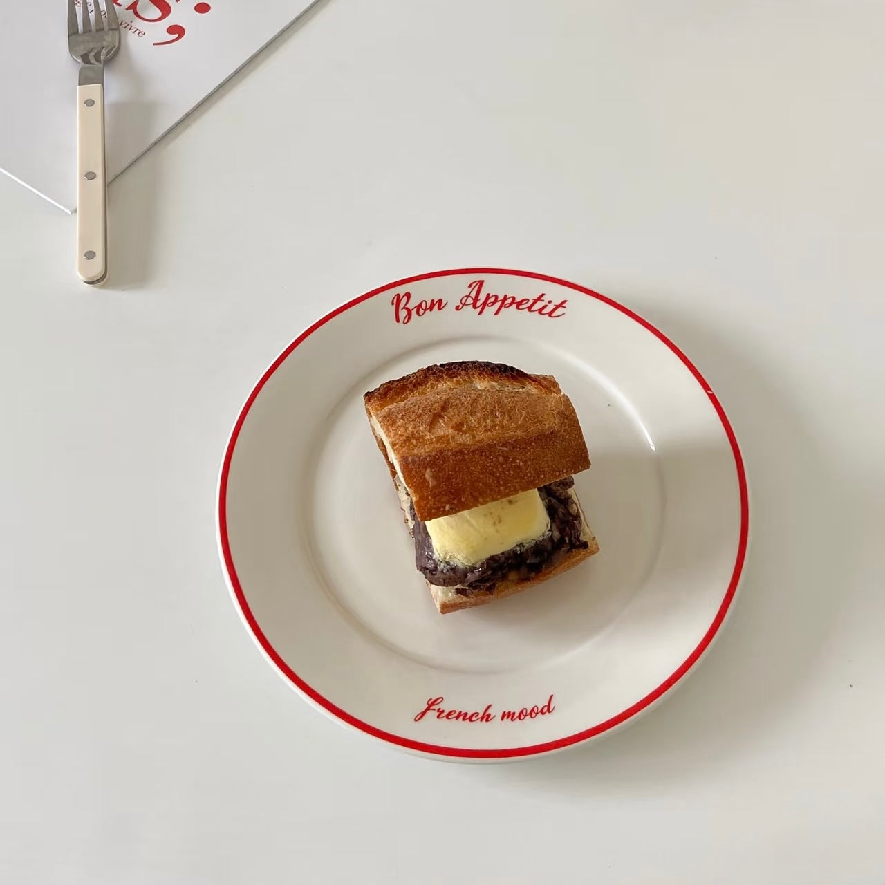 A small sandwich on a white ceramic plate with a red rim and the words 'Bon Appetit' and 'French mood' printed on it, photographed on a white tabletop with a fork partially visible at the top left.