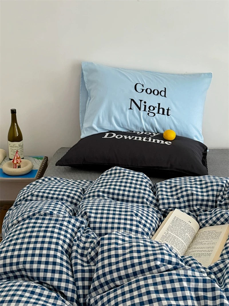 Bed with checkered blue and white bedding, pillows with 'Good Night' and 'Happy Downtime' text, and a bottle on a nightstand.
