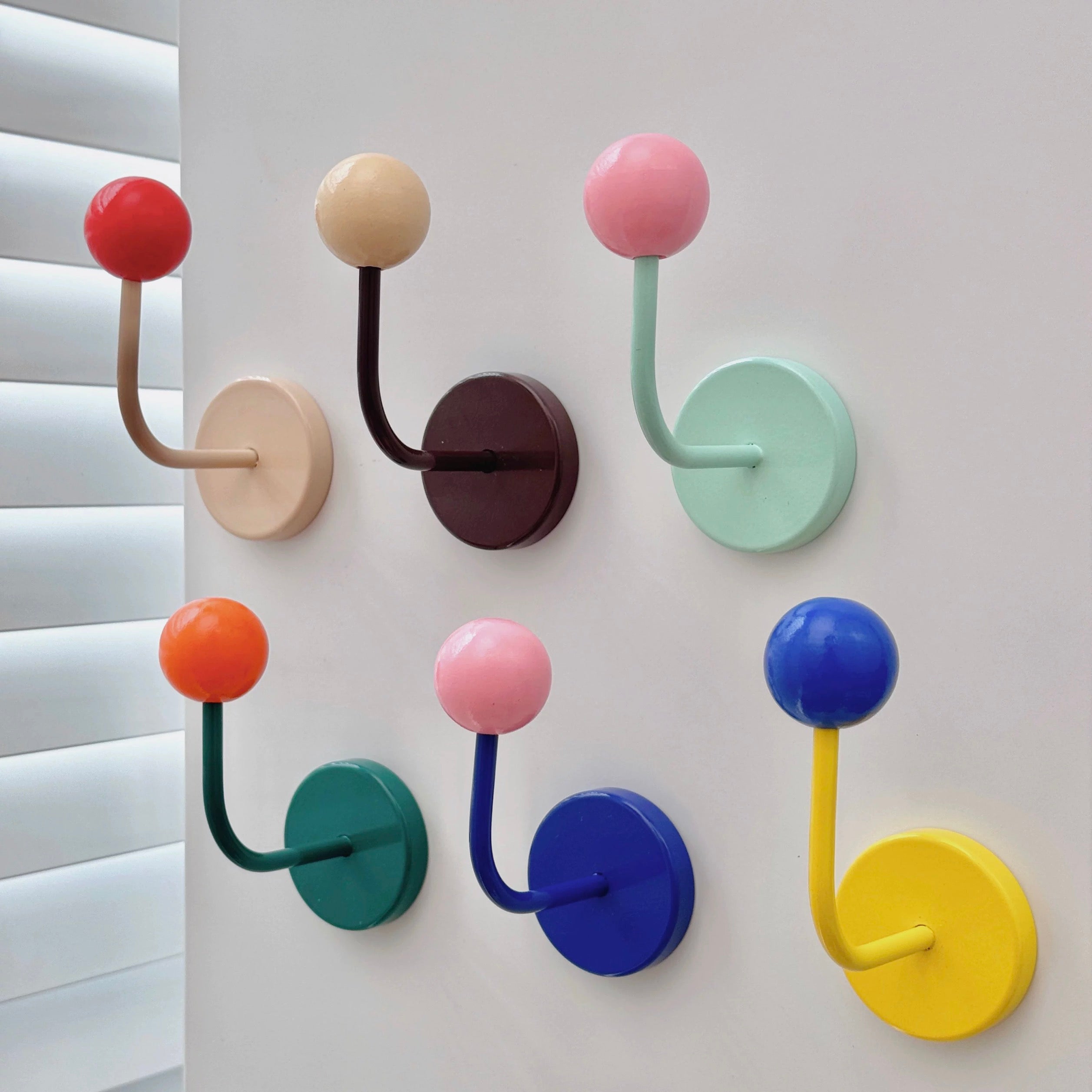 Six wall-mounted coat racks in assorted colors, including red, beige, dark brown, mint green, orange, pink, teal, blue, and yellow, arranged on a white wall next to window blinds.