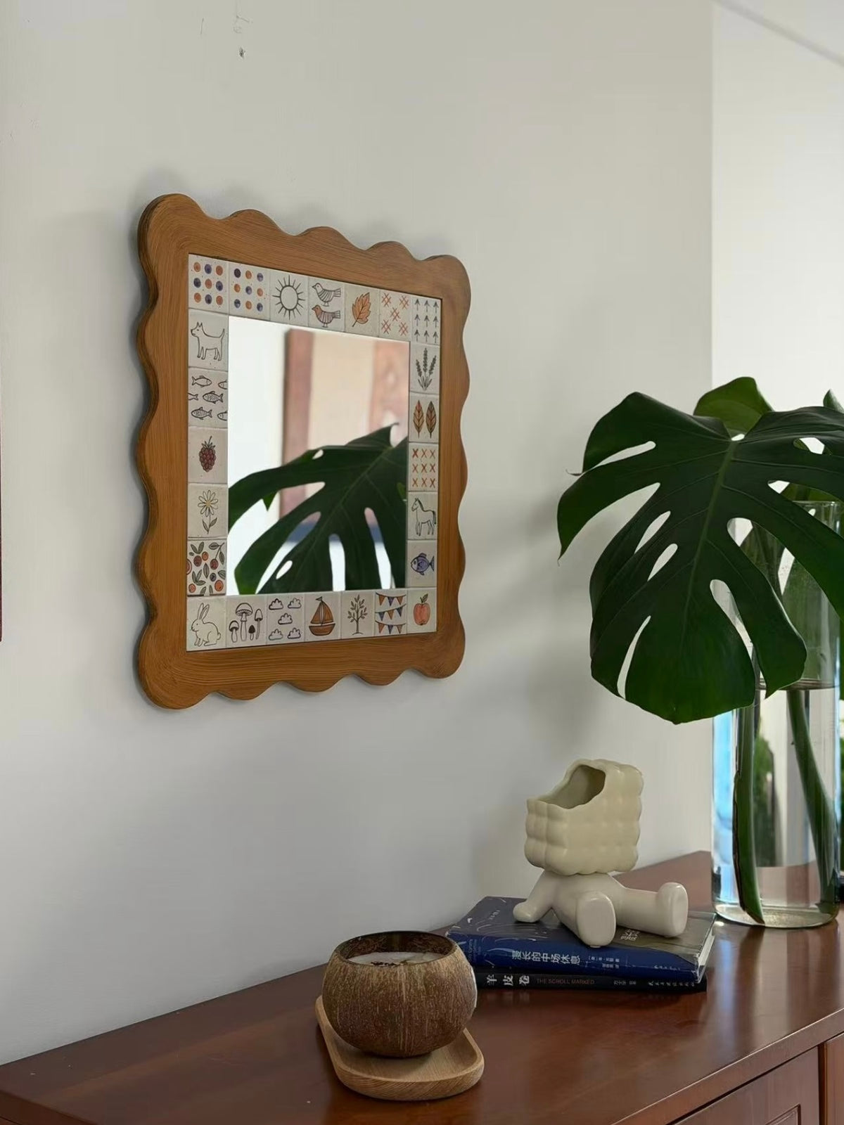 A brown wall mirror with an illustrated border hangs beside a large green leaf above a cabinet.