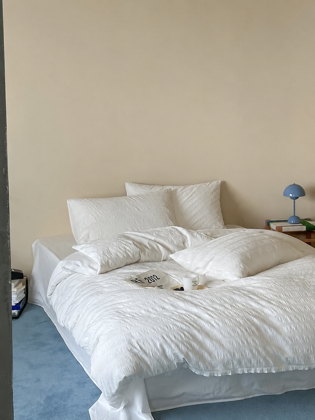 A bed dressed in white textured bedding, including a duvet cover and multiple pillows, set against a plain beige wall with a small blue bedside lamp on a wooden nightstand.