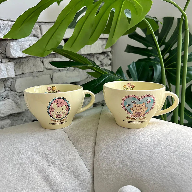Two pale yellow ceramic cups with cute cartoon animal illustrations, displayed on a white shelf next to a white ribbed vase.