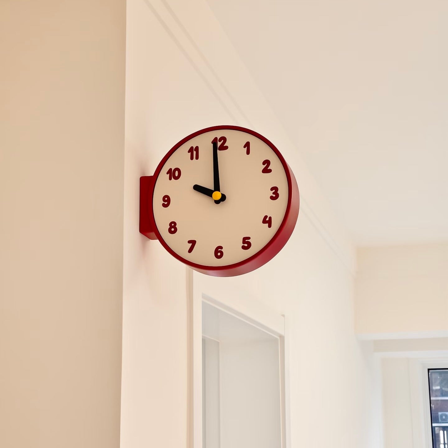 A round wall clock with a dark red frame and cream-colored face, mounted on a beige wall in a hallway.