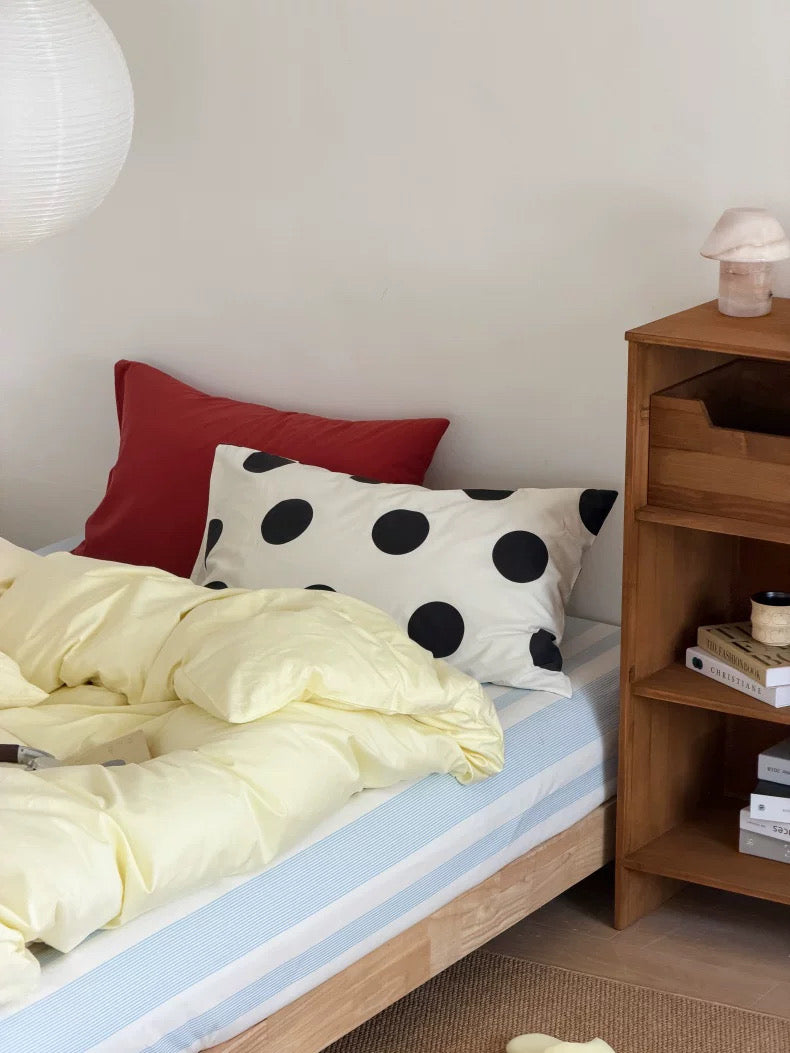 A cozy bedroom scene with a bed made up in pale yellow bedding, a light blue and white striped sheet, a red pillow, and a white pillow with large black polka dots. A wooden nightstand sits beside the bed against a light wall.